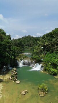 Vertical drone ascends and pulls back from Pangas Falls, showing cascades, green pool, rocks, palms, and hills in Bilar