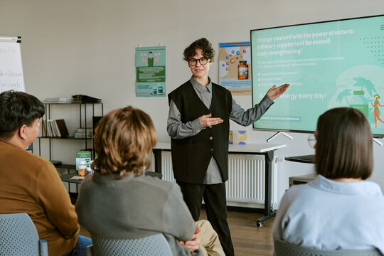 Young trainer presenting supplement product information to business audience during workshop, explaining wellness benefits and sales messaging. Useful for corporate training or health marketing