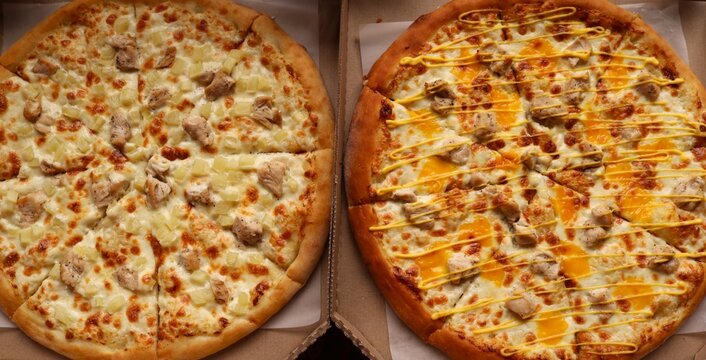 Two round pizzas in cardboard boxes. On the left is a classic Hawaiian pizza with chicken and pineapple pieces on mozzarella cheese. On the right is a cheese pizza with chicken, drizzled with sause