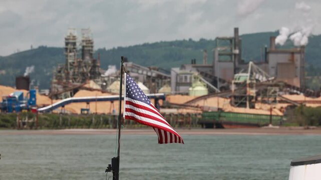 American flag waves above water near industrial pier, hazy pulp mill complex with silos plus conveyor towers lining Longview, Washington river waterfront under gray sky. Aerial drone view.