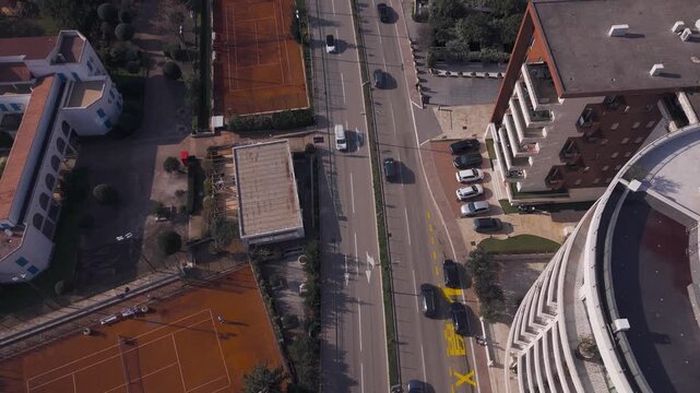 Cinematic drone flyover parallel to a busy two-way traffic road, tilting up to reveal a city panorama with a park, tennis court and mountains.