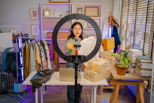Young Asian woman fashion entrepreneur live streaming selling clothes online with smartphone ring light and laptop in home studio, representing small business owner and professional women.