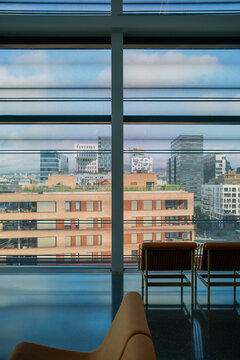 Near the window, a chair anchors the foreground as reflection opens through glass, revealing an interior city view where daylight shapes distant architecture