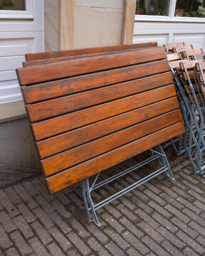 Wooden tables lean against a building outside a cafe in a city during the day with some chairs stacked nearby