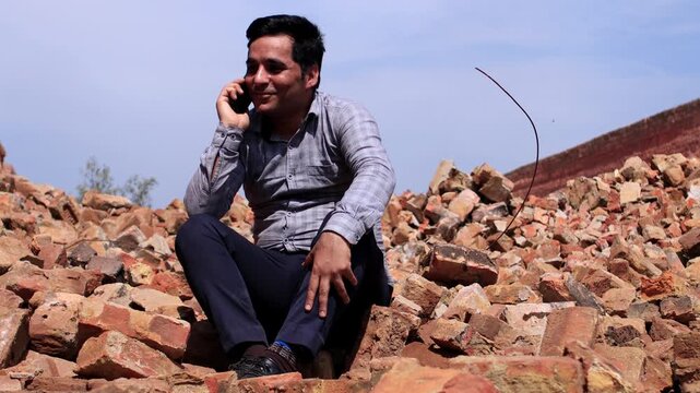 Cheerful young men of Indian ethnicity sitting near brick manufacturing factory and using mobile phone video portrait.