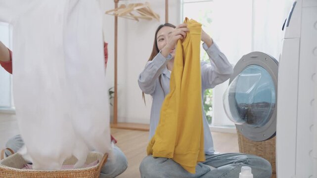 Two happy and cheerful Asian women friends sitting on floor, unloading freshly washed clothes from washing machine or dryer and folding together with smiles in white minimal, cozy laundry room at home