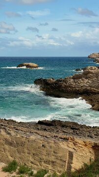 Crystal clear Mediterranean waves crashing against rocky shoreline near Cala Agulla, Mallorca. Vertical coastal landscape with modern seaside hotels, dramatic natural scenery. 