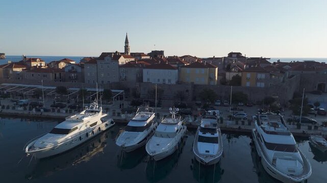 Cinematic drone flyover above the promenade and yachts, revealing a close-up view of Budva Old Town main bell tower with the Adriatic sea and St. Nicholas island in the background, Montenegro.