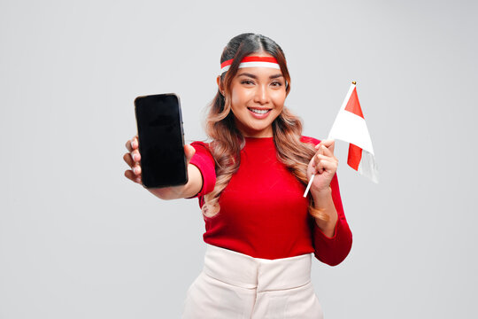 Cheerful Indonesian woman holding a mini country flag while showing copy space on her phone screen.