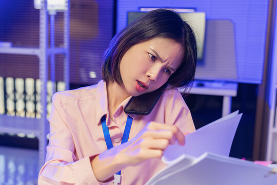 Young Asian businesswoman talking on smartphone while reviewing documents in office.