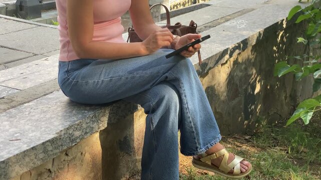 A relaxed woman sits on a stone ledge holding a smartphone. She peacefully checks her phone while enjoying the quiet outdoor greenery. Her comfortable posture reflects calm contentment.