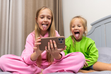 Two young girls making funny faces while watching something on a smartphone at home © Алина Бузунова