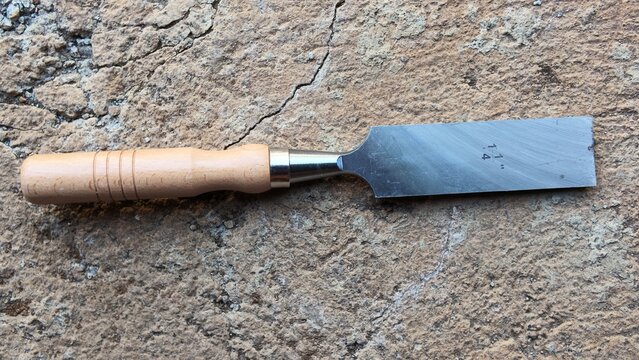 A wide flat wood chisel tool with a wooden handle lying flat against a textured concrete surface.
