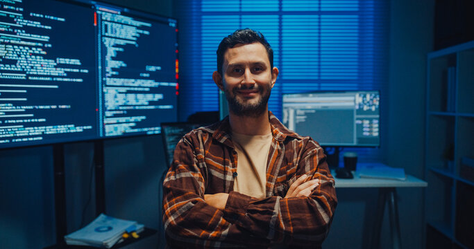 Portrait of confident Caucasian male programmer smiles looking at camera in office at night. Professional developer workspace, Secure computer network system, Advanced cybersecurity data concept.