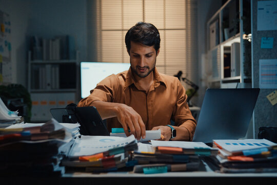 Overworked young Latin businessman working late on document in large stack of paperwork files at office night. Tax audit deadline, accounting stress, corporate burnout, urgent business concept.