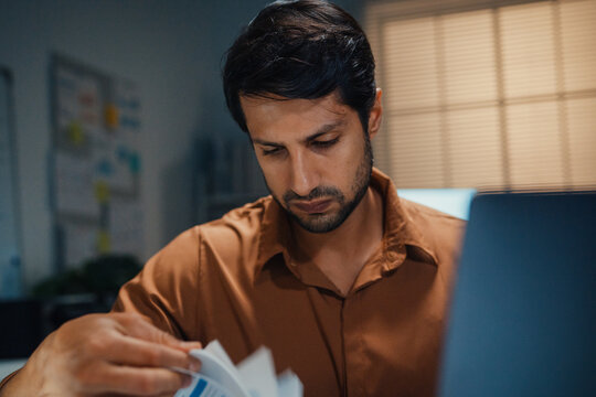 Close up overworked young Latin businessman working late on document in large stack of paper files at office night. Tax audit deadline, accounting stress, corporate burnout, urgent business concept.