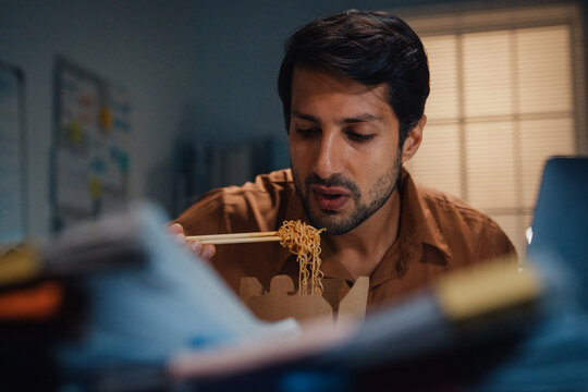 Young Latin businessman eating takeout instant noodles and reviewing document on laptop at desk in dark night office. Overtime working late, overworked deadline, fast food, corporate problem concept.