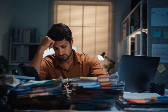Stressed young Latin businessman having severe headache and migraine at desk in night office. Debt crisis, corporate burnout, late overtime workload, mental health anxiety, business failure concept.