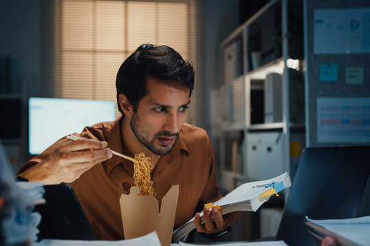 Young Latin businessman eating takeout instant noodles and reviewing document on laptop at desk in dark night office. Overtime working late, overworked deadline, fast food, corporate problem concept.