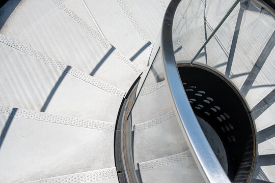 An overhead view follows a spiral staircase, where a railing circles the steps as layered shadows turn interior architecture into sweeping curves and geometry across the frame