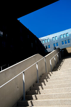 A concrete staircase rises beside a dark building as the railing guides perspective over the steps, while shadow cuts through exterior architecture beneath blue sky