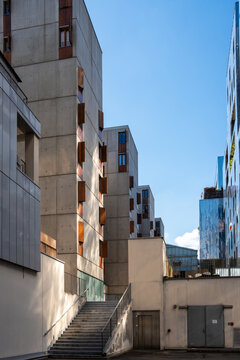 Within a narrow urban courtyard, perspective leads toward a broad concrete staircase between buildings, as exterior facades frame blue sky above the layered architecture