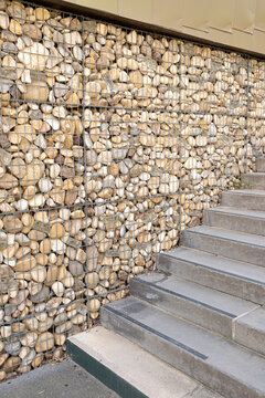 Measured perspective follows a concrete staircase beside a gabion retaining wall, where packed stones shape the steps and tactile texture of the exterior architecture