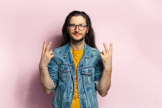 Young confident man with long hair showing peace gesture isolated on pastel pink background.