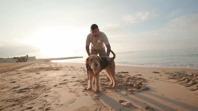 Man walking his airedale terrier dog on a sandy beach at sunrise