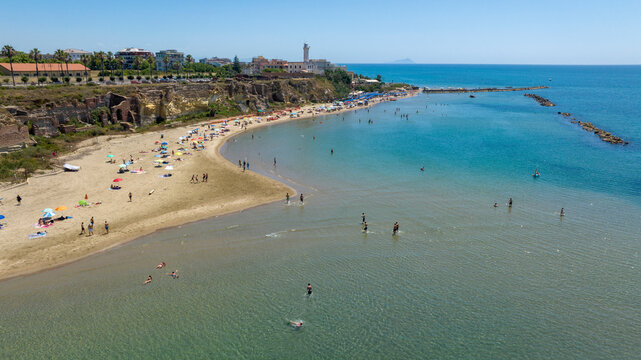 Aerial view of a sandy bay with clear waters where people are swimming, sheltered by stone breakwaters under a bright blue sky, with a coastal cliff and buildings of Anzio, Italy, in the background.