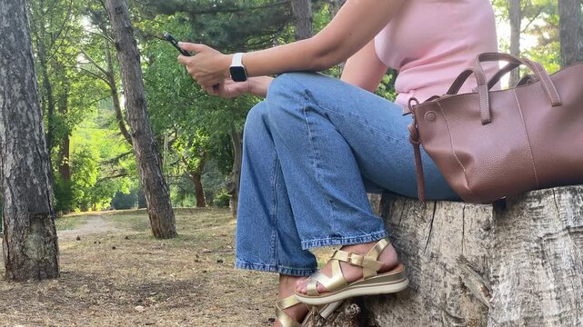 A relaxed woman sits on a tree stump using her phone. She rests peacefully in a quiet, sunlit park. Her posture shows calm contentment.