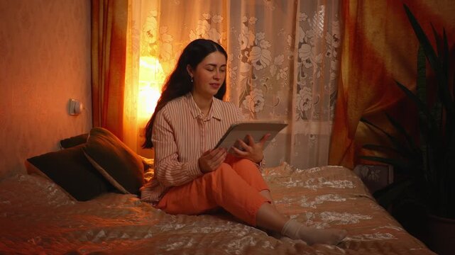 Beautiful young Caucasian woman with braces relaxing and smiling while watching an online video on a tablet in the evening. Digital entertainment, home leisure, orthodontic treatment, and modern lifes