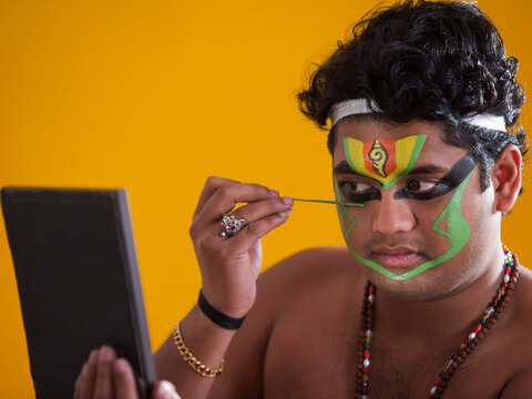 Kathakali dancer looking int the mirror and applying green colour on his face. 