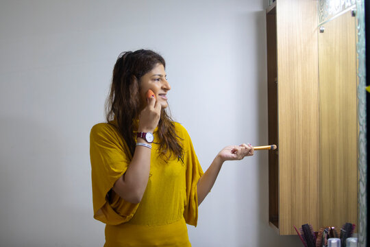 Woman looking in the mirror and blending her makeup while holding a makeup brush in her other hand.