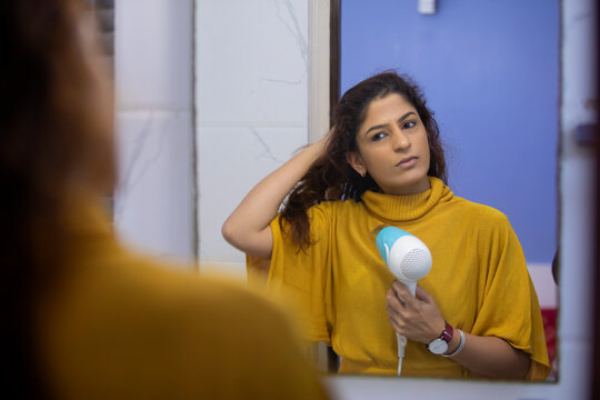 Woman drying her wet hair with a hairdryer while looking in the mirror in her bathroom. 