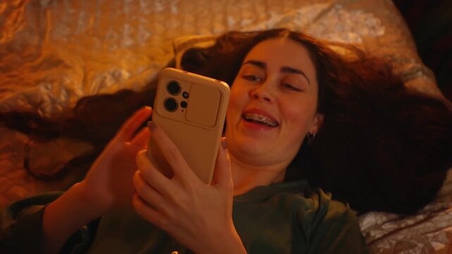 Top view of a young Caucasian woman with long hair lying on a bed and using a smartphone with a surprised happy expression. Digital communication, good news, online entertainment, and modern lifestyle