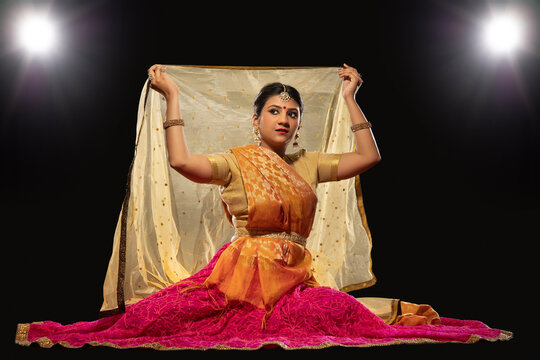 Kathak dancer sitting gracefully and performing in front of a dark background. 