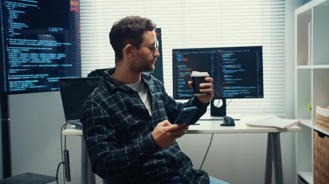 Young Caucasian male software engineer enjoy coffee break with smartphone during work at desk. Workday productivity and concentration, Freelance career lifestyle, Modern tech office workplace concept.