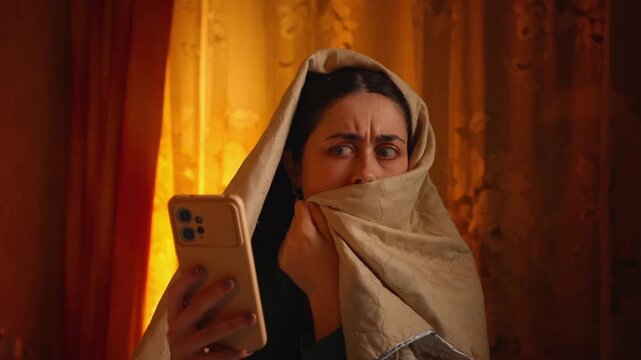 Portrait of a frightened Caucasian woman wrapped in a blanket using a smartphone while reading bad news online. Shock, fear, anxiety, and emotional reaction concept
