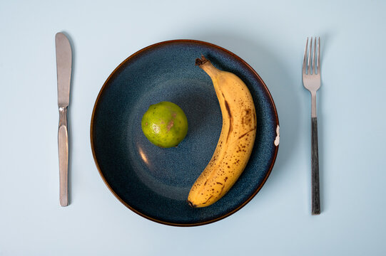 Imperfect lime and ribe banana on a dented blue plate with worn mismatched cutlery, representing anti-consumerism, valuing functional objects and using what we already have instead of replacing it.