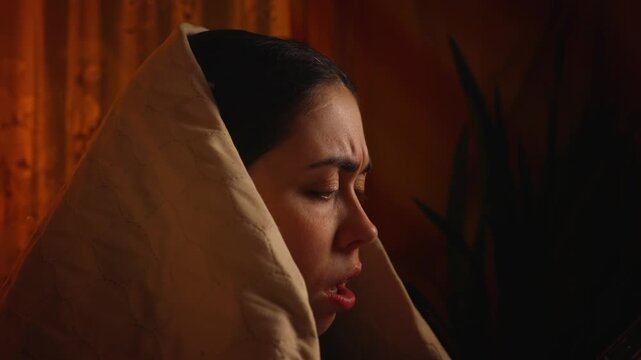 Side view close up of a frightened woman wrapped in a blanket using a smartphone while discovering bad news online. Shock, fear, anxiety, and emotional reaction concept.