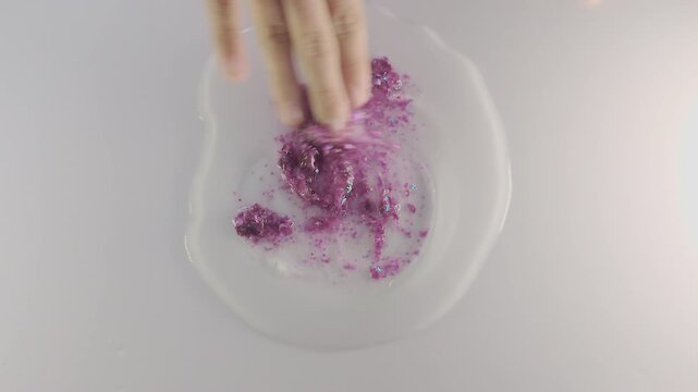 A close-up, top-down view captures a child's hand mixing shiny pink glitter and sequins into a puddle of clear slime on a white tabletop surface during a popular home science STEM experiment.