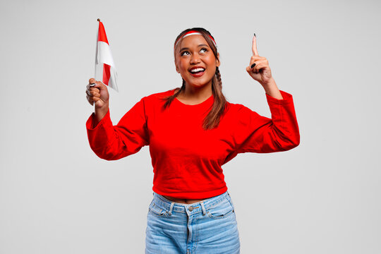 Joyful young Asian woman is holding mini country flag and pointing to the copy space upwards.