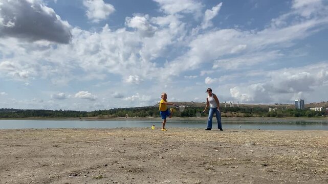 A happy boy plays with a racket while a woman walks nearby on a lakeside shore. They enjoy a fun day outdoors under a cloudy sky. The playful game brings pure joy and warmth.