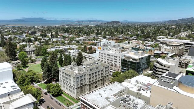 Cinematic drone pullback over Berkeley, California, featuring the downtown commercial district, office and retail buildings, dense urban development, and scenic hills in the background on a bright sun