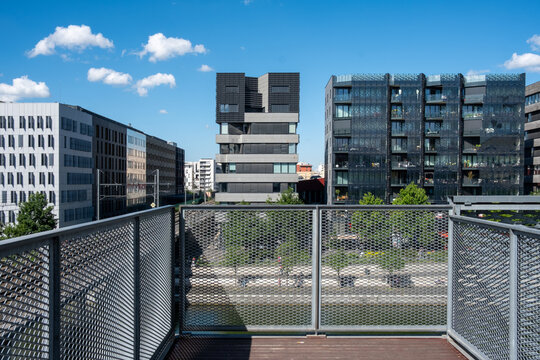 A gated walkway and metal railing lead toward urban buildings in La Confluence, Lyon, France, creating a layered perspective through contemporary architecture and open sky