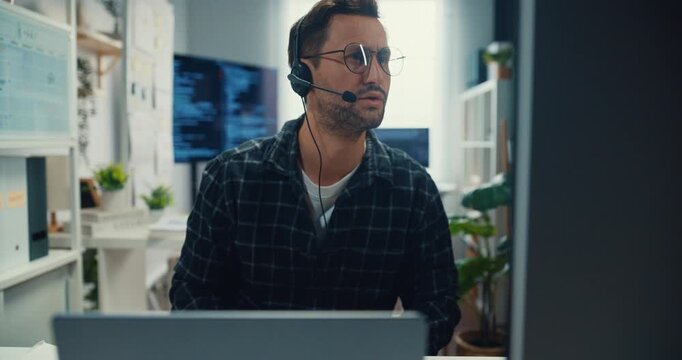 Young Caucasian male technical support specialist wearing headset speaks on computer at office. Professional call center help desk, Cyber security operator service, Online client consultation concept.