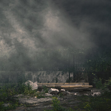A gloomy, desolate landscape featuring heavy smoke rising over a concrete wall, overgrown vegetation, and abandoned debris under a dark sky. Halloween and horror background