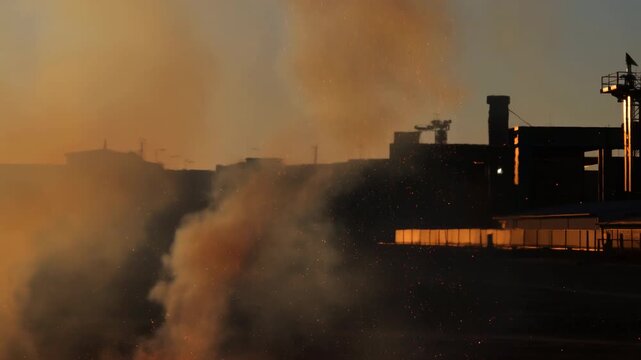 expanding flames and smoke in an industrial fire at a factory or complex, dramatic and urgent scene viewed from the side of the burning buildings with visible chimneys and concrete andmetal.