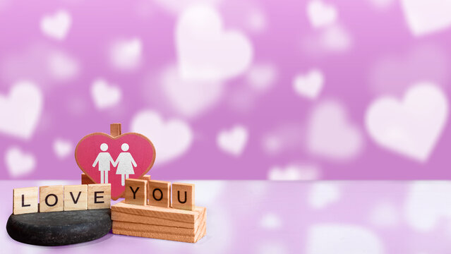 Wooden letter blocks spelling 'LOVE YOU' with a couple heart decoration on a soft purple background featuring heart-shaped bokeh. Concept of First Love Day